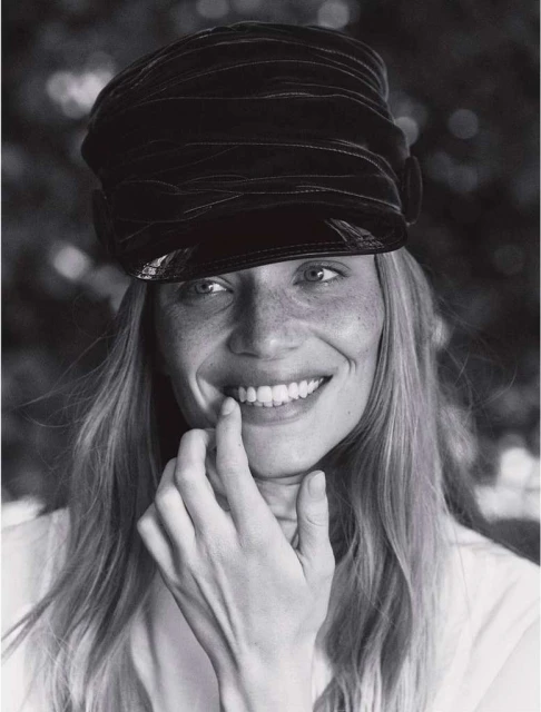 Black and white portrait of a smiling woman with blonde hair wearing a black hat, with her fingers touching her lips.