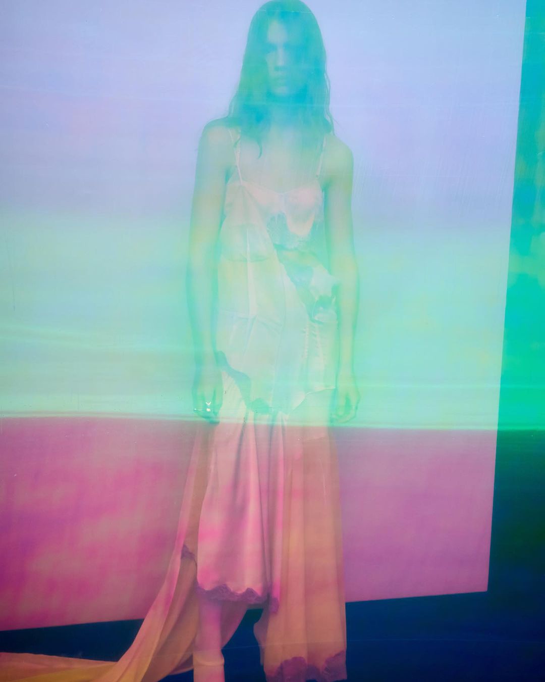 Woman in sheer dress poses in a studio with soft, diffused lighting and a split, pastel-colored backdrop; long exposure blurs details.