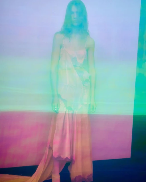 Woman in sheer dress poses in a studio with soft, diffused lighting and a split, pastel-colored backdrop; long exposure blurs details.