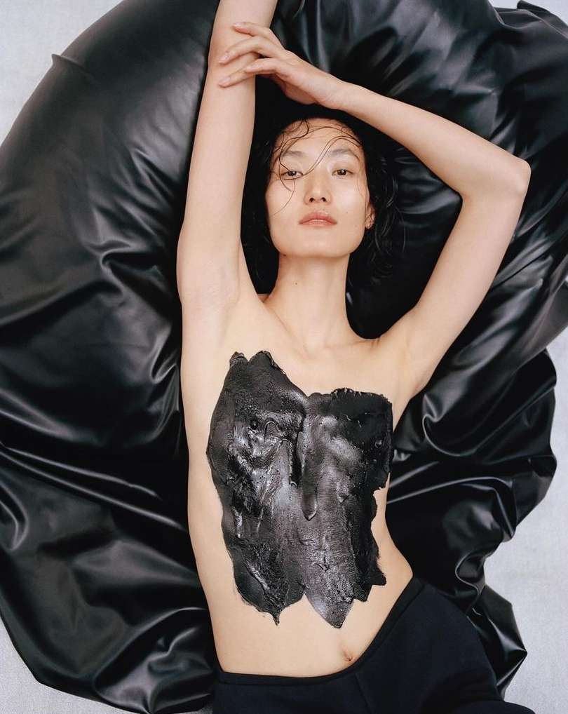 Asian woman lies on a shiny black material with black substance on her chest. Arms are raised and hair is wet. Indoor, neutral background.