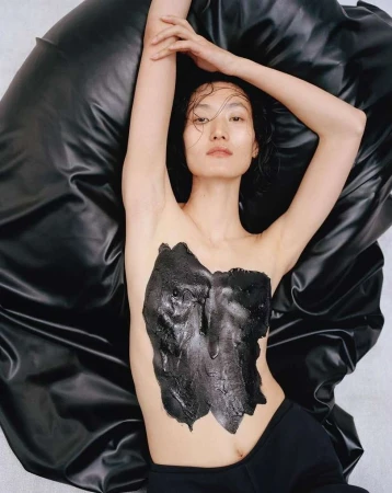 Asian woman lies on a shiny black material with black substance on her chest. Arms are raised and hair is wet. Indoor, neutral background.