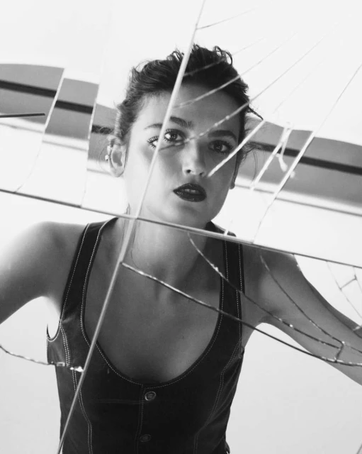 Black and white portrait of a woman with dark hair in a denim top, looking through cracked glass fragments in a studio setting.