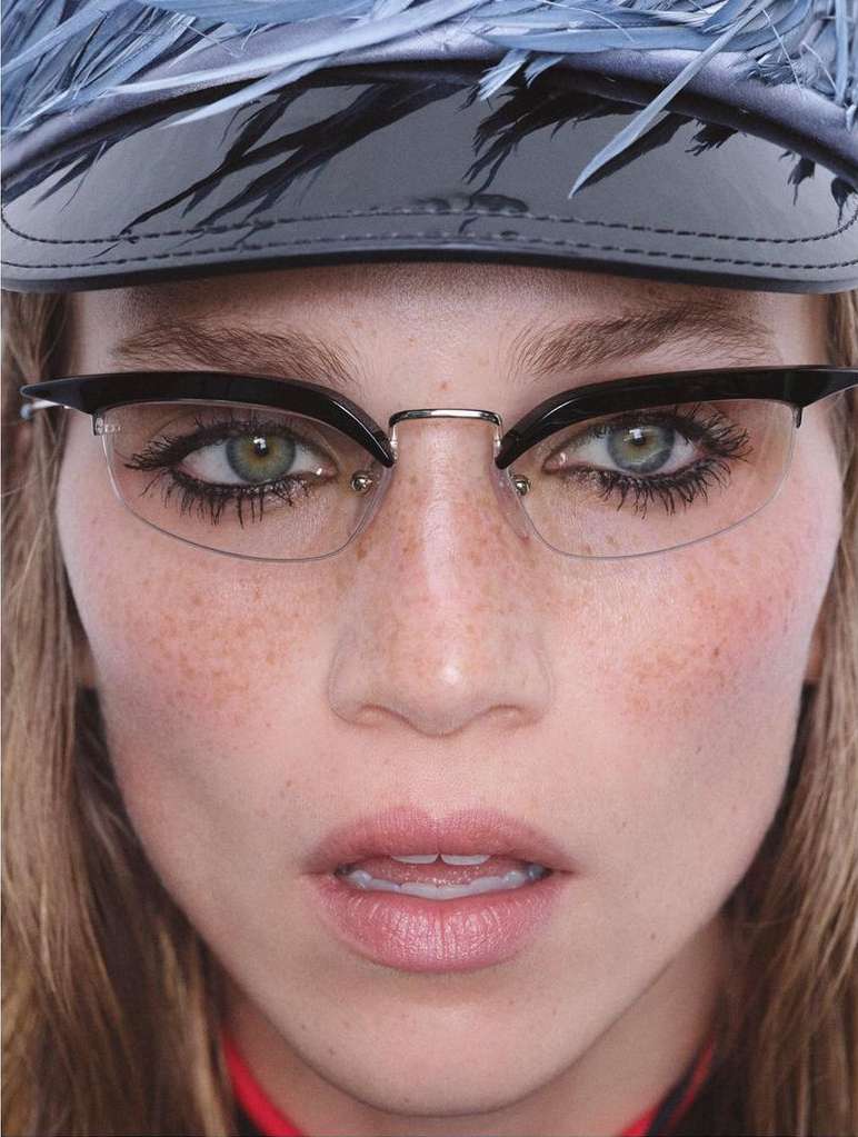 Close-up of a woman with freckles wearing black-and-silver framed glasses and a feathered hat looking directly at the camera.
