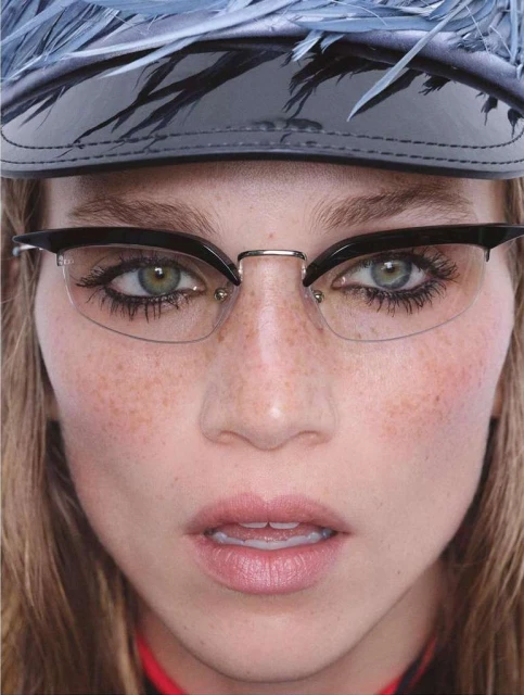 Close-up of a woman with freckles wearing black-and-silver framed glasses and a feathered hat looking directly at the camera. 