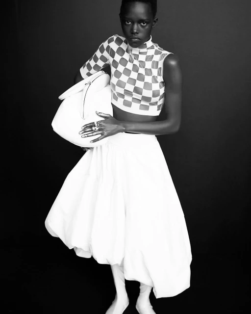 Monochrome portrait of a Black woman wearing a checkered crop top and a full white skirt, holding a white purse against a dark backdrop.