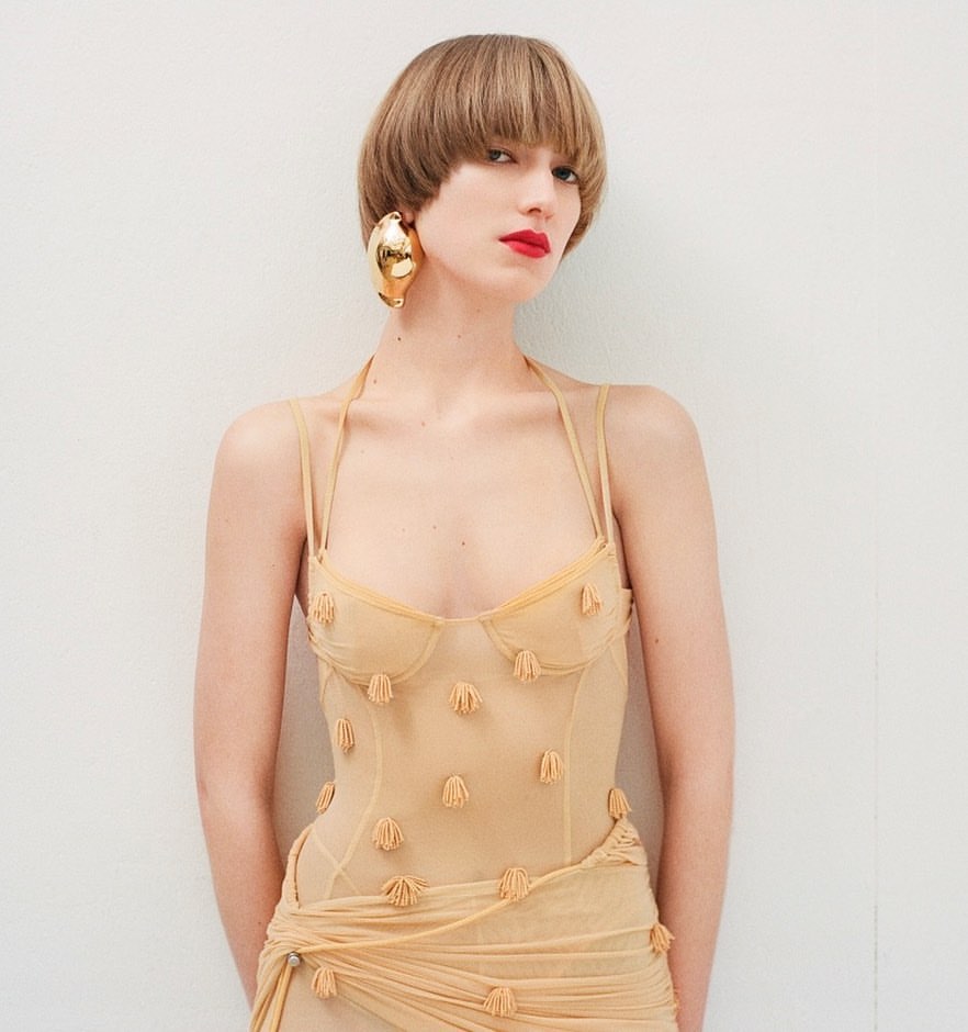 Woman in sheer dress with sculptural earrings poses against a white wall in a studio, fashion photography.