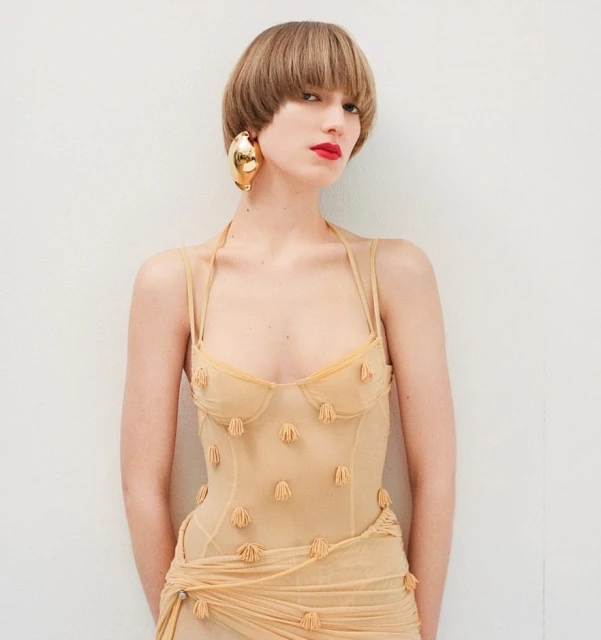 Woman in sheer dress with sculptural earrings poses against a white wall in a studio, fashion photography.