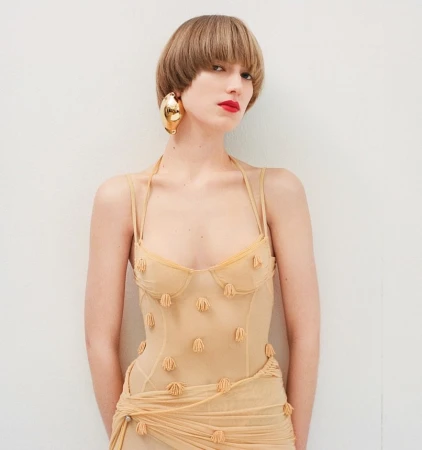 Woman in sheer dress with sculptural earrings poses against a white wall in a studio, fashion photography.