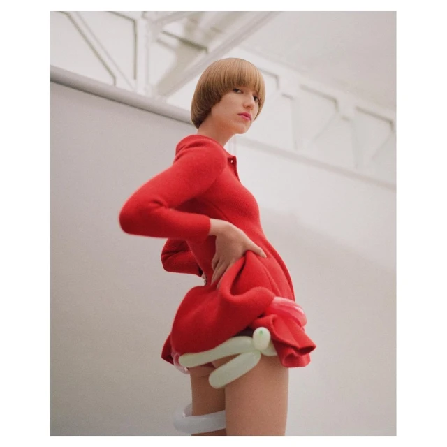 A woman with a blonde bowl cut poses in a red dress with balloon details, in an indoor studio with white walls and overhead lighting.