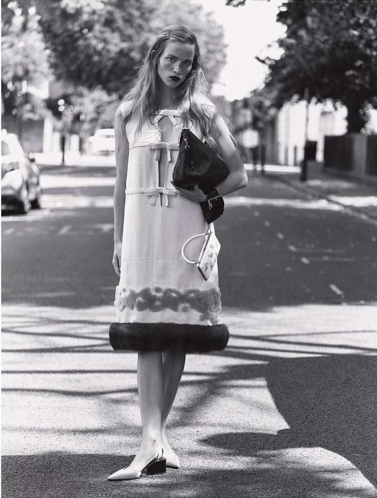 A full shot in black and white of a woman wearing a dress and carrying a handbag, standing in the middle of a street with cars parked along the side.