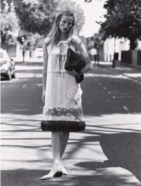 A full shot in black and white of a woman wearing a dress and carrying a handbag, standing in the middle of a street with cars parked along the side.