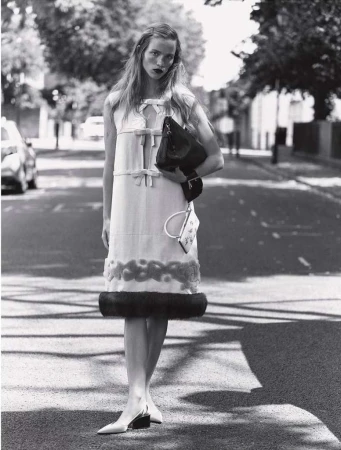 A full shot in black and white of a woman wearing a dress and carrying a handbag, standing in the middle of a street with cars parked along the side.
