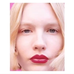 Close-up beauty portrait of a blonde woman with glossy crimson red lips and subtle pink eye makeup against a vibrant pink background.