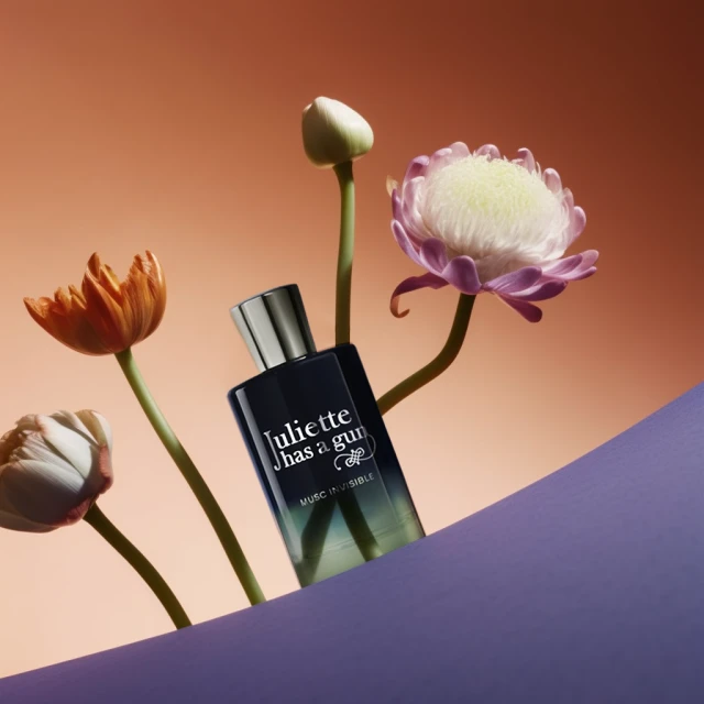 Scent bottle by Juliette Has A Gun surrounded by colorful flower blossoms and partially obscured by purple paper. Perfume still life.