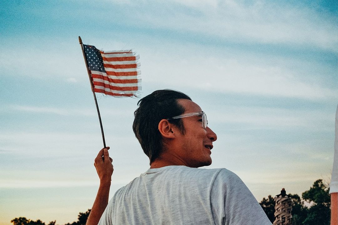 Adult male gazes off into distance while holding a tattered United States flag against a serene blue and white sky at dusk.