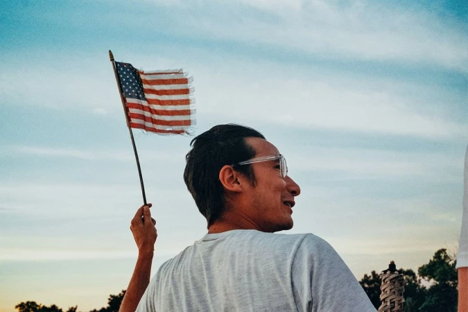 Adult male gazes off into distance while holding a tattered United States flag against a serene blue and white sky at dusk.