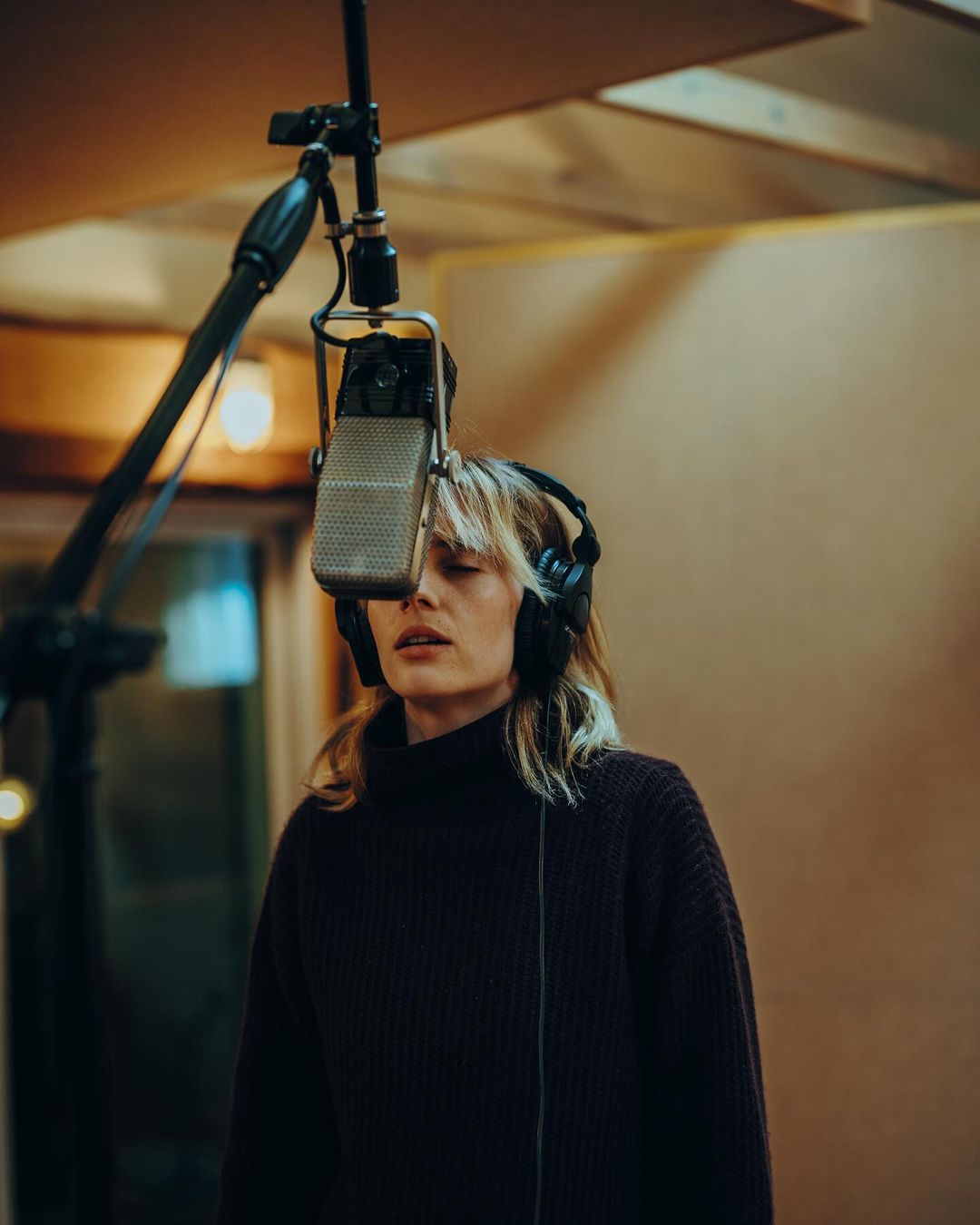 A woman in a dark turtleneck sweater sings into a vintage microphone in a recording studio, wearing headphones with soft, diffused lighting.