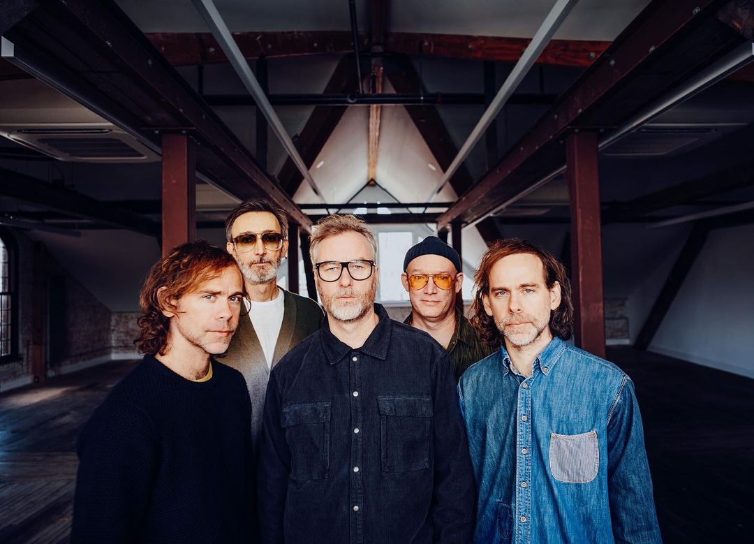 Indoor portrait of a musical group of five adult males, wearing casual clothing, posing in a room with a high ceiling and beams.