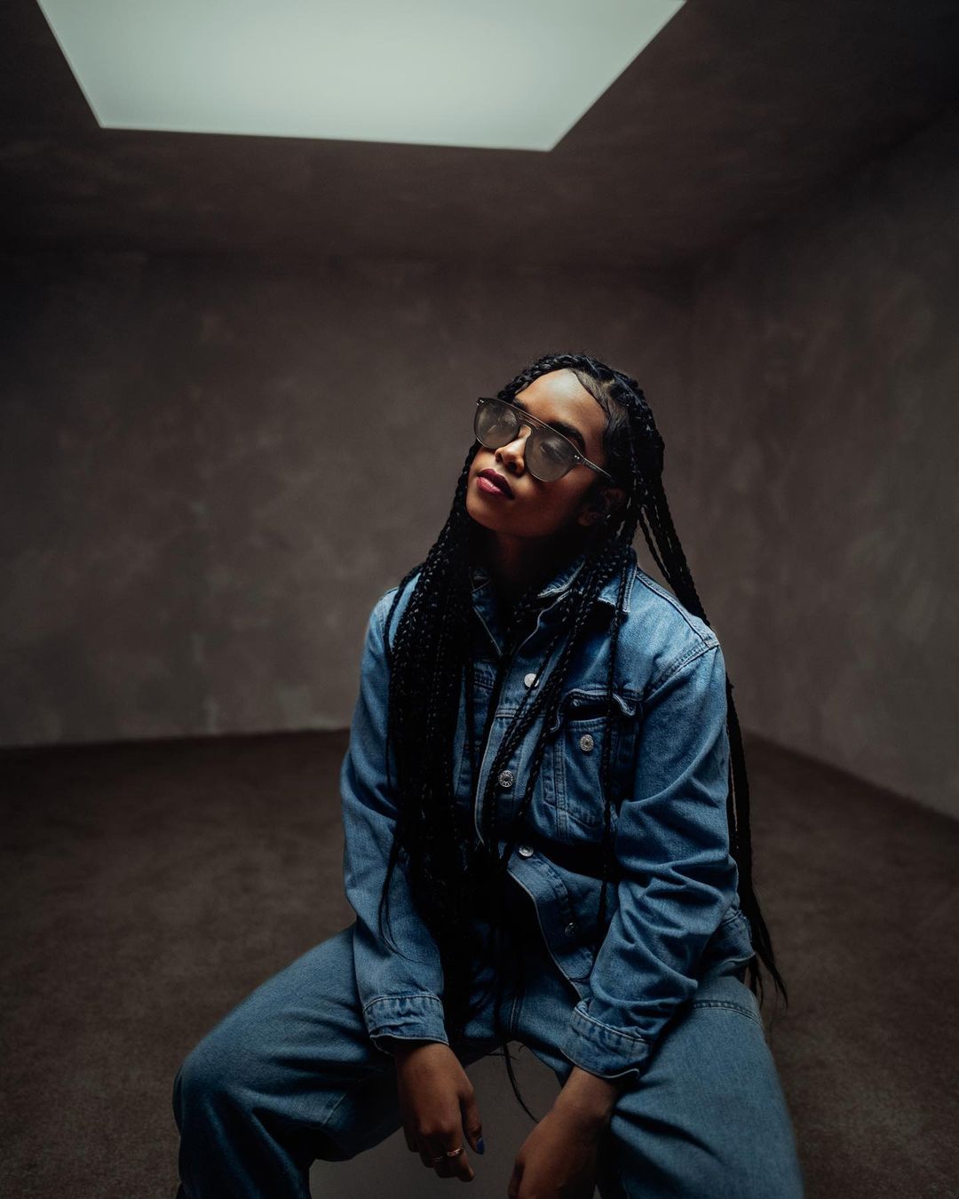 Woman with braided hair poses in a denim outfit with sunglasses in a room with soft lighting from an above window.