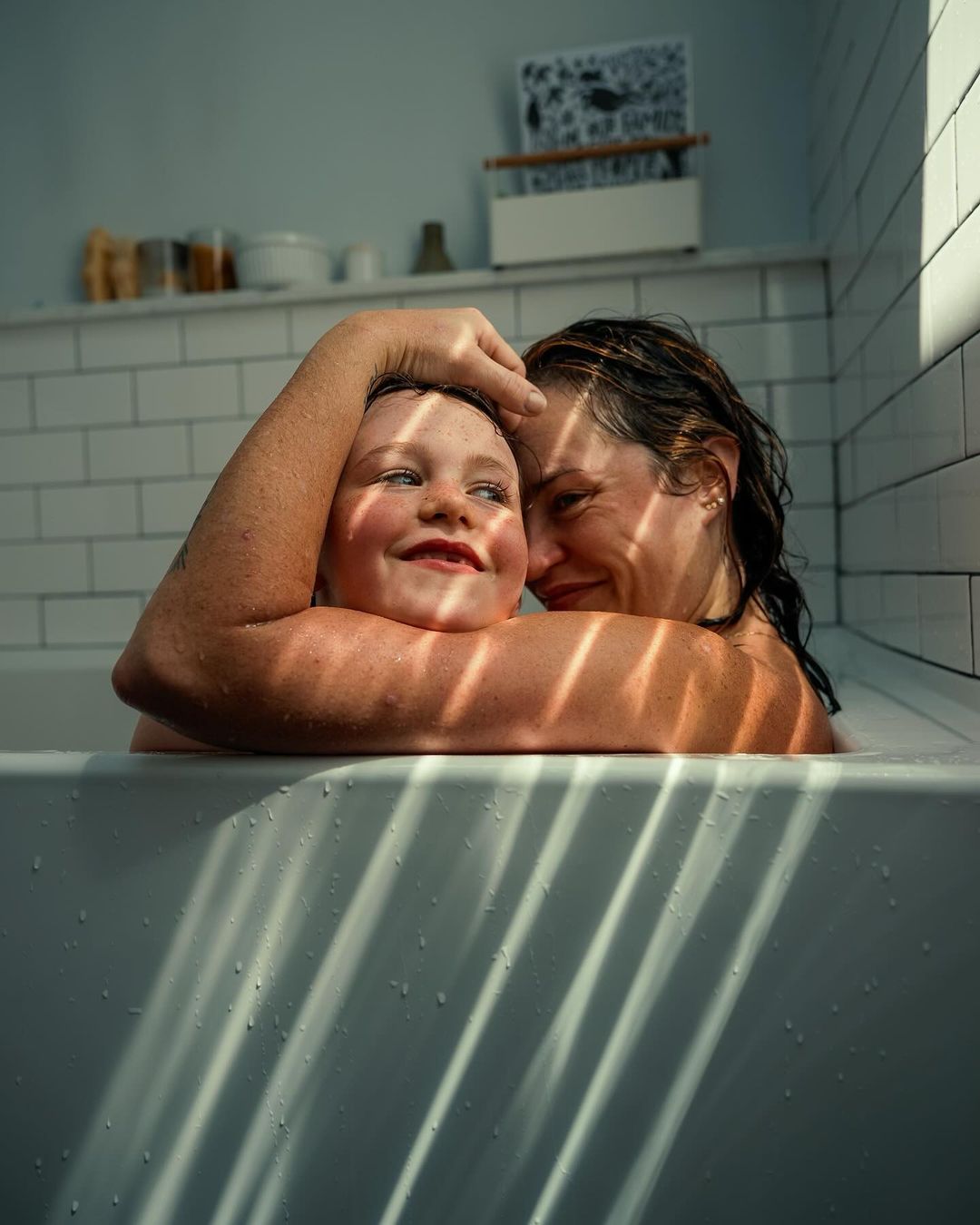 A smiling woman and a young child are embraced in a bright, naturally lit white-tiled bathroom with sunlight creating linear shadows across them.