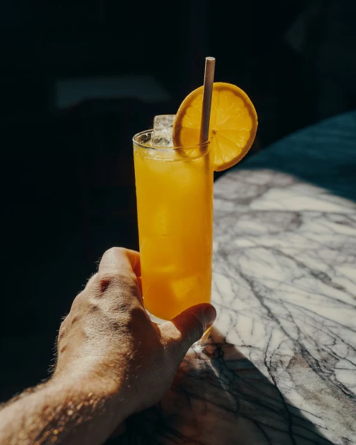 Refreshing orange beverage with ice and a straw, held by a hand, set on a marble surface with shadows.