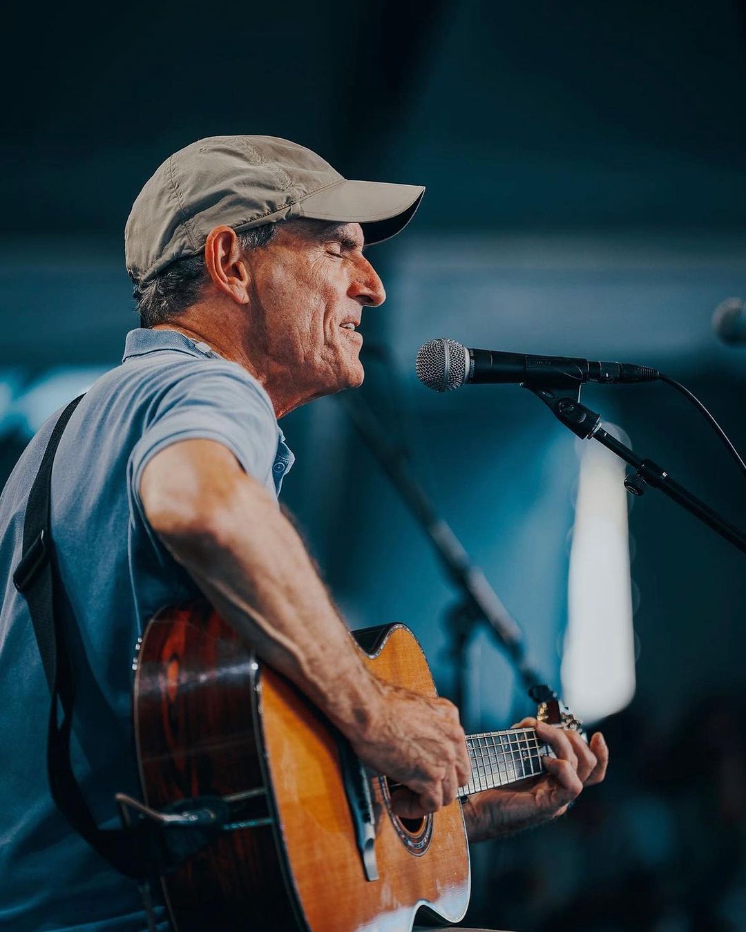 Adult man playing an acoustic guitar and singing into a microphone on stage, wearing a cap and casual clothing in an indoor setting.
