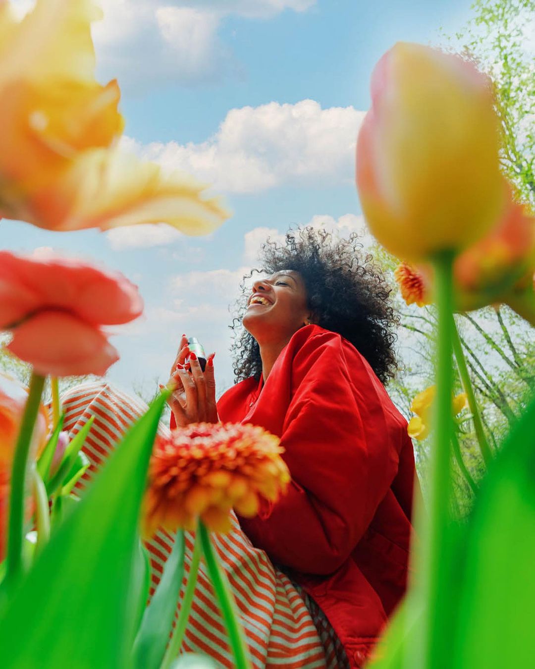 Woman with curly hair in a red jacket, sitting among colorful tulips under a blue sky with clouds, looking up and smiling in natural light.