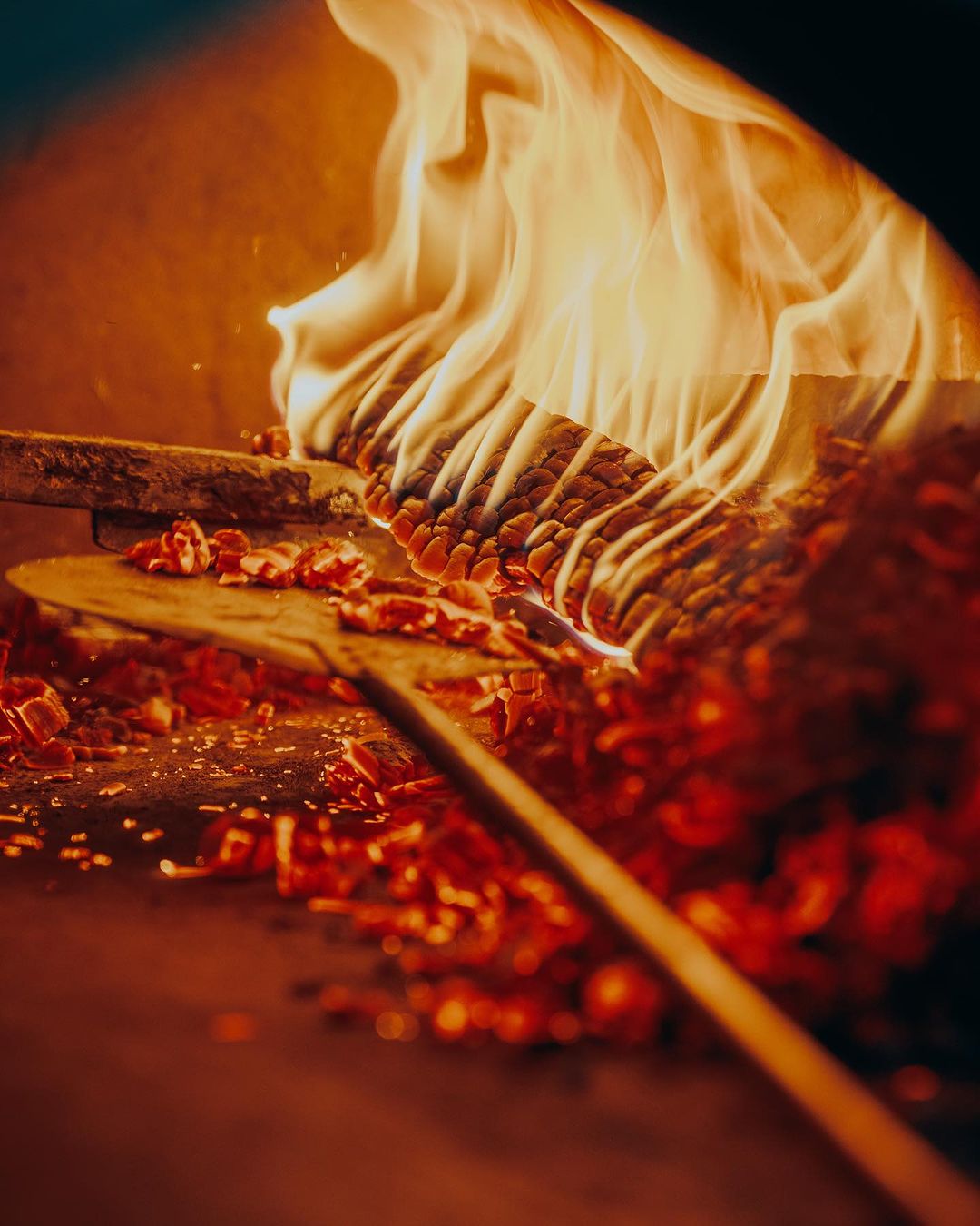 Close-up of flames engulfing wood in a pizza oven, with a pizza peel and embers creating a warm, inviting atmosphere.