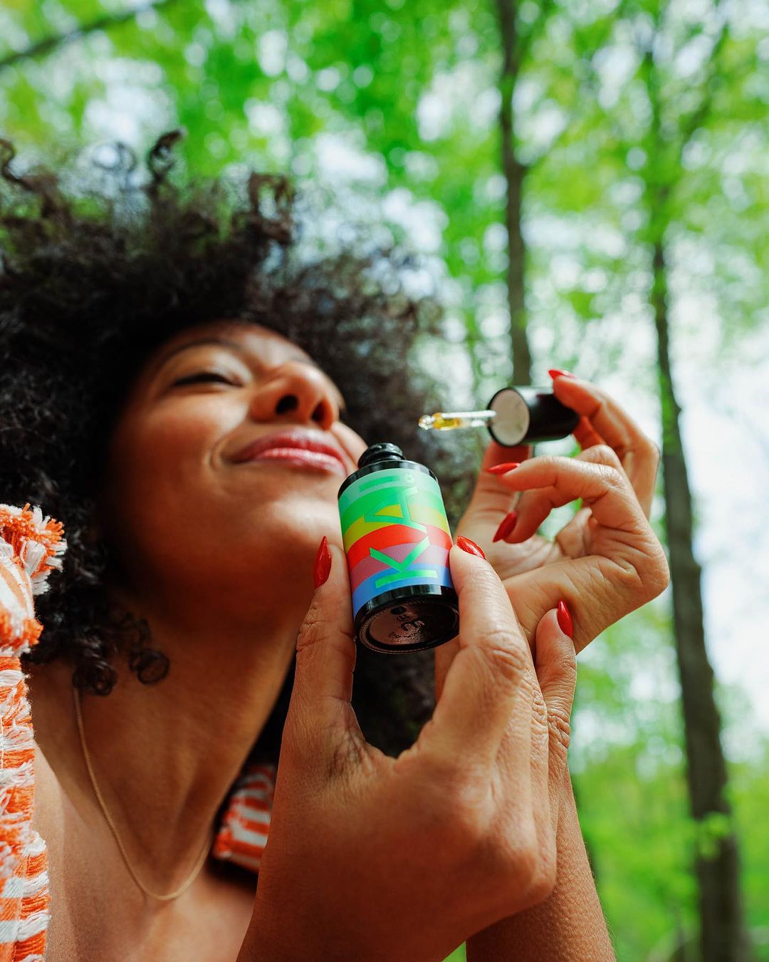 Close-up shot of a woman with curly hair holding a colorful skincare serum bottle with a dropper in a forest setting.