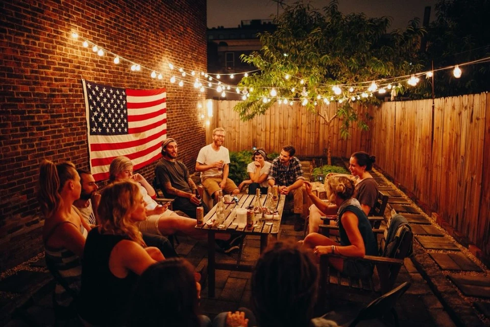 Group of ten people gather outdoors at dusk with string lights and American flag on brick wall, creating a warm, convivial ambiance.