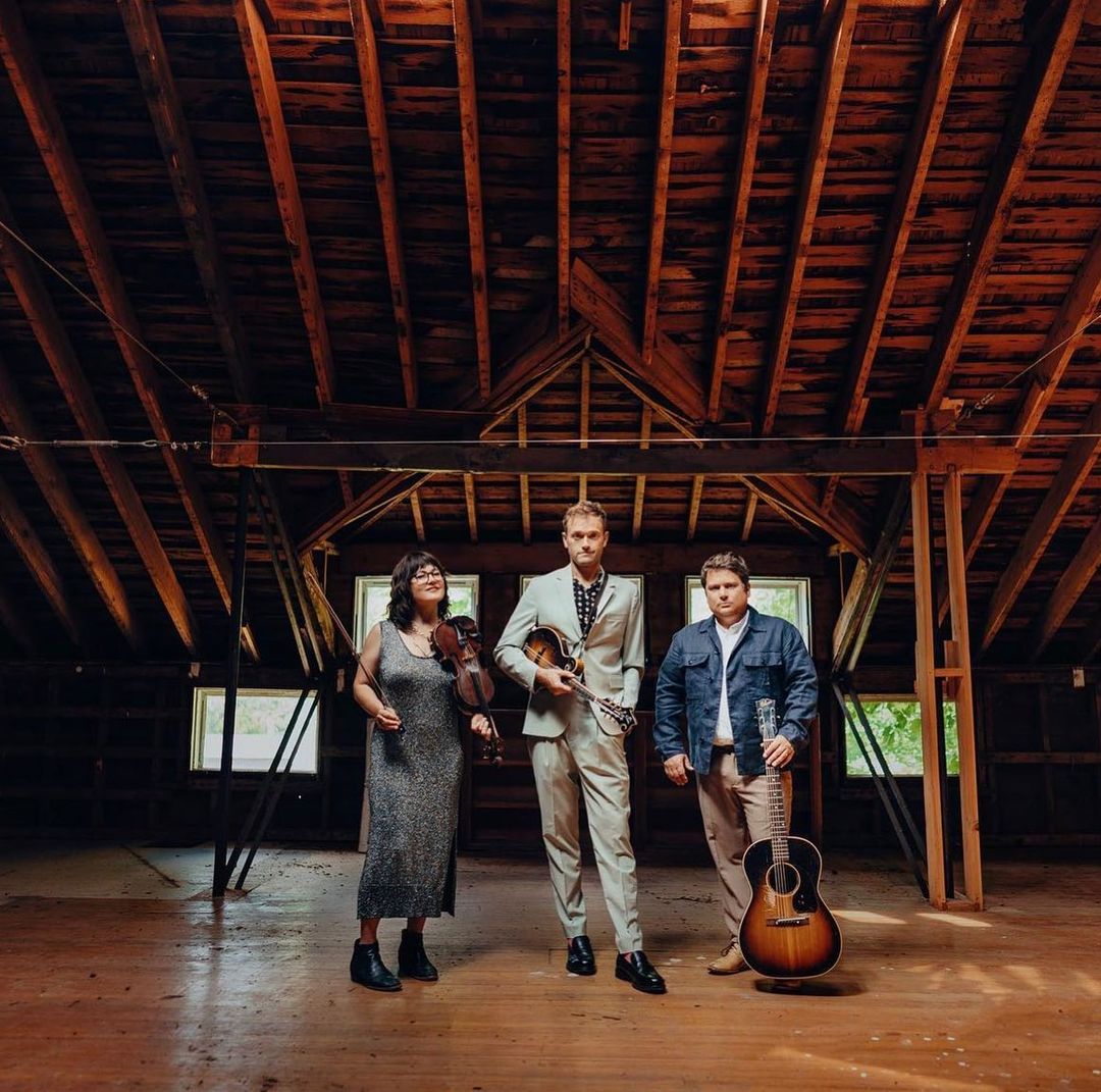 Three musicians in a rustic attic studio. A woman holds a violin, a man holds a mandolin, and a man stands with a guitar.