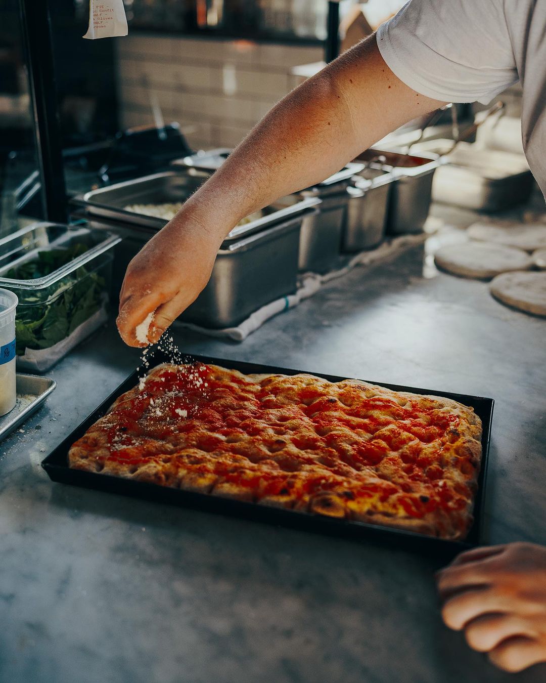 Rectangular pan pizza being sprinkled with cheese by a chef in a restaurant kitchen setting; close-up view with food preparation.