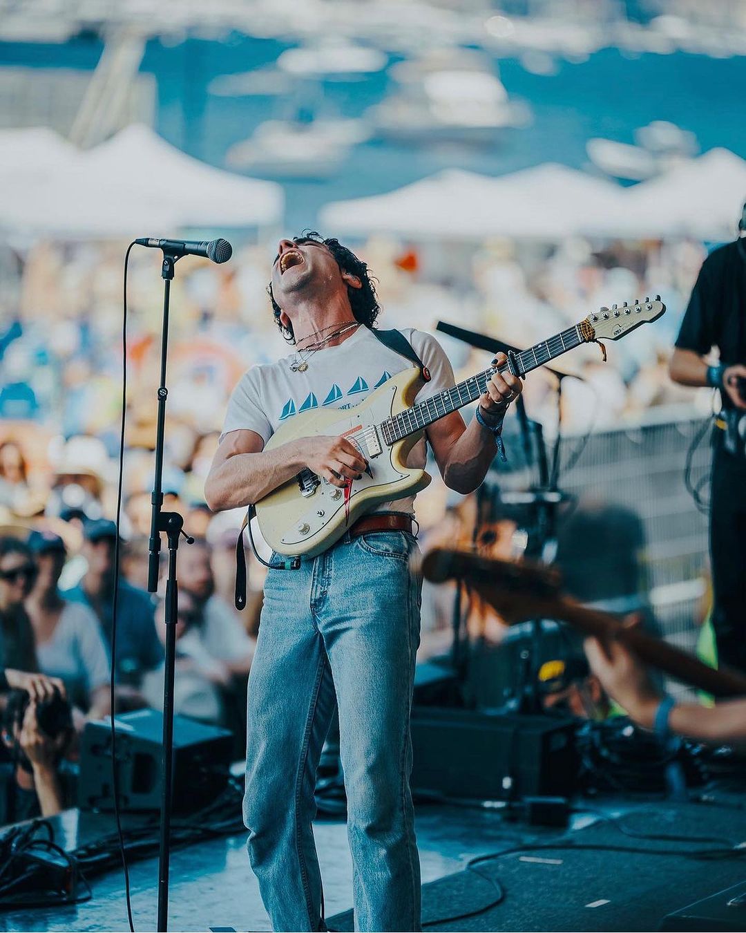 Male musician passionately performing on stage, playing electric guitar into a microphone with a crowd visible in the background.