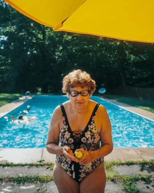 Elderly woman in a floral swimsuit and goggles holds a yellow ball near an outdoor swimming pool with a yellow umbrella overhead.