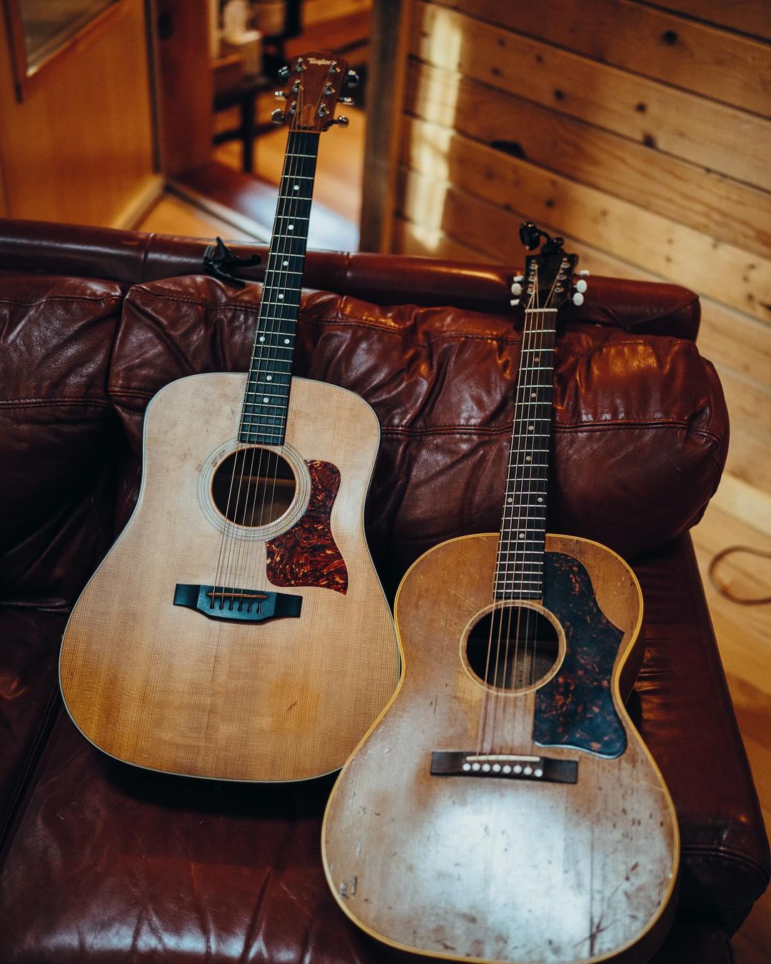 Two acoustic guitars lean against a brown leather couch in a room with wood-paneled walls, bathed in soft, warm light.