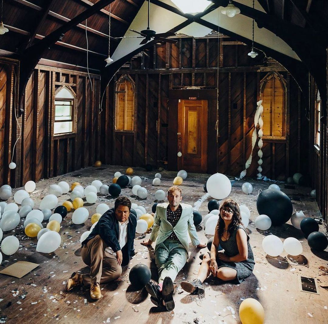 Three adults and a dog sit among white, gold, and black balloons on a weathered wood floor in a rustic, bare-walled room with arched windows.