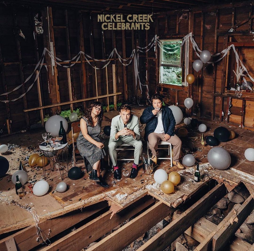Music album cover featuring three individuals seated amidst balloons and debris in a dilapidated room, promoting the band Nickel Creek.