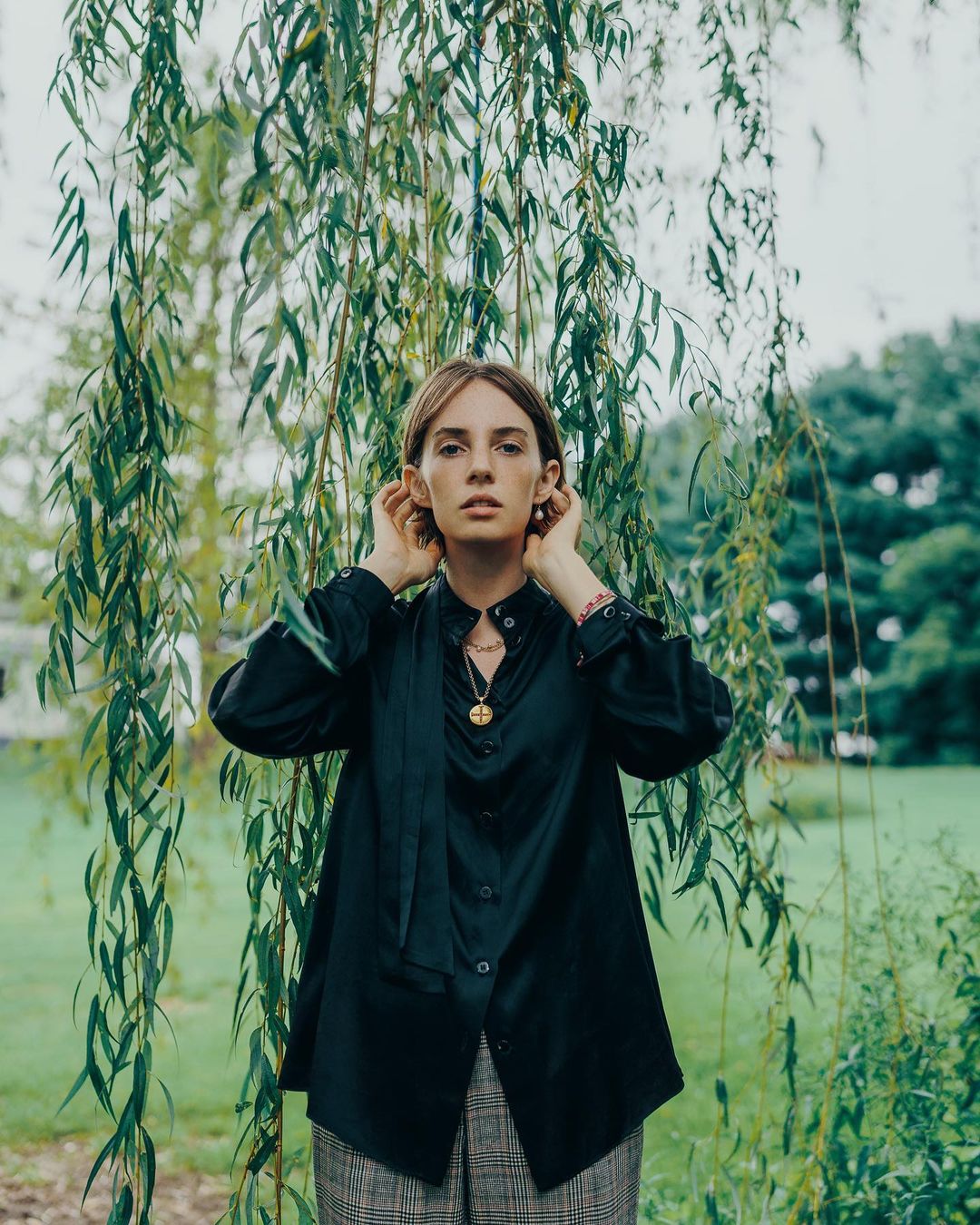 Young woman poses in black shirt and plaid pants under a willow tree with green foliage in an outdoor setting on a cloudy day.