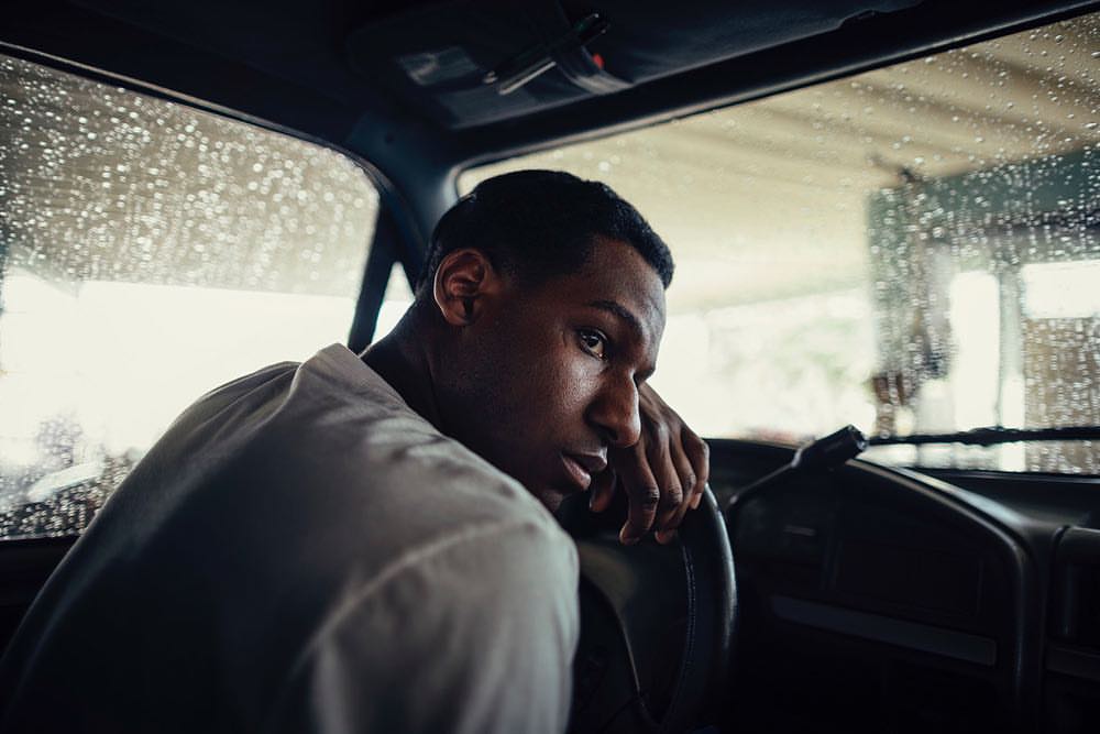 Man in a car during a rain, glancing back over his shoulder with a white collared shirt and dark hair.