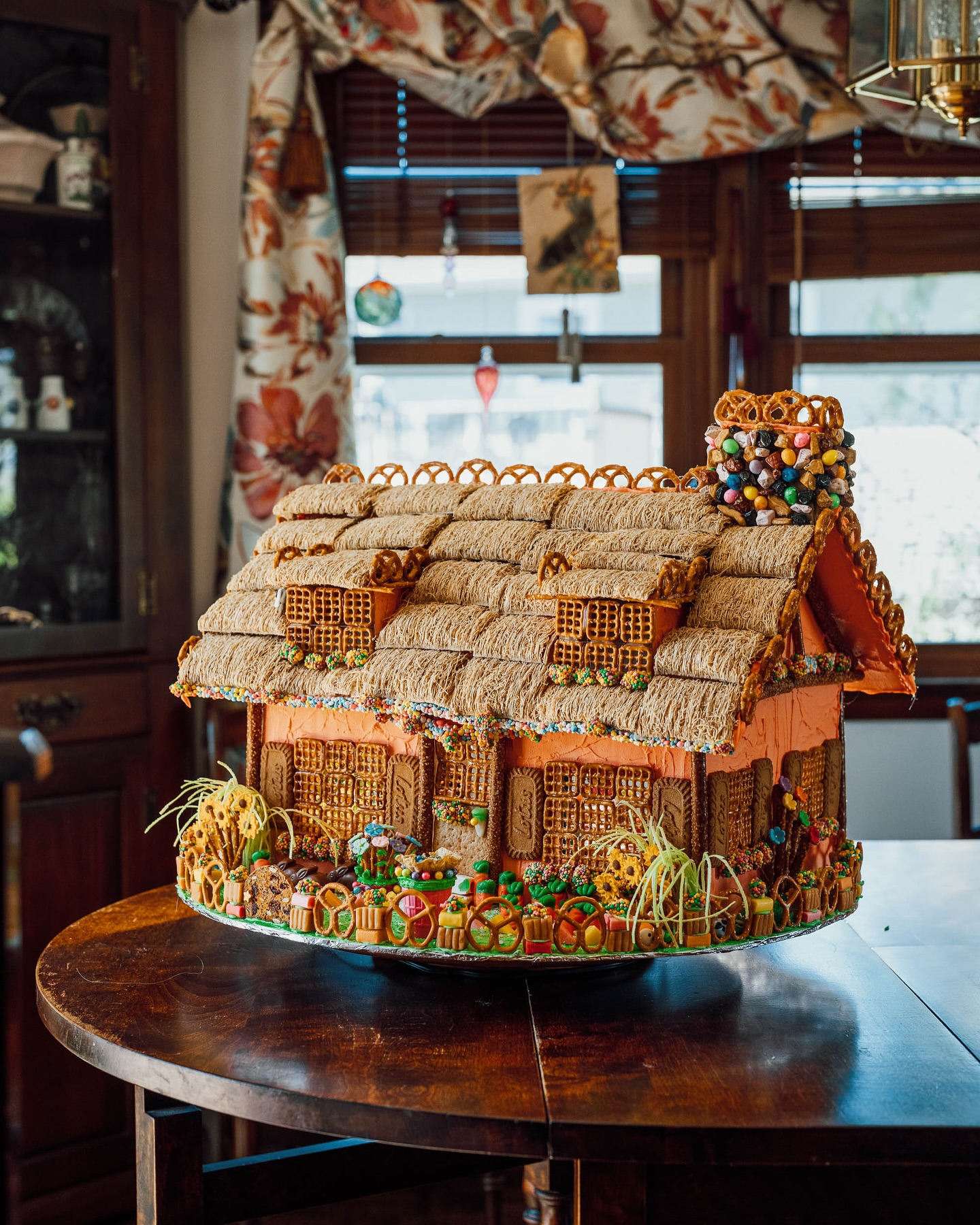 Whimsical gingerbread house decorated with candy and pretzels, sitting on a wooden table in front of a window with floral curtains.