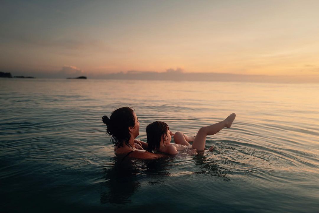 A mother holds a child in the ocean water at sunset. The sky is a blend of orange and pink hues. The water is calm and reflective.