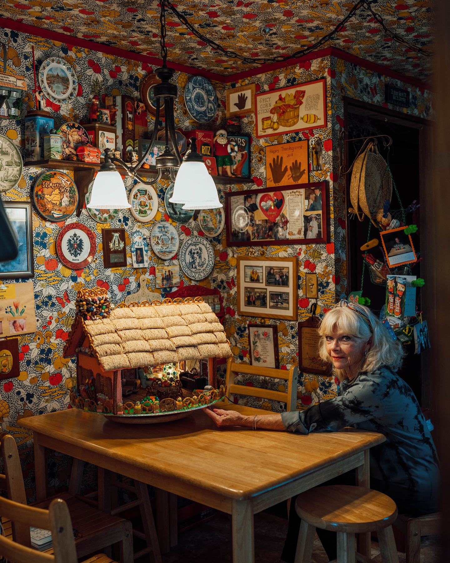 Elderly woman poses at wooden table with craft house model in kitsch, decorated room with patterned wallpaper and hanging plates.