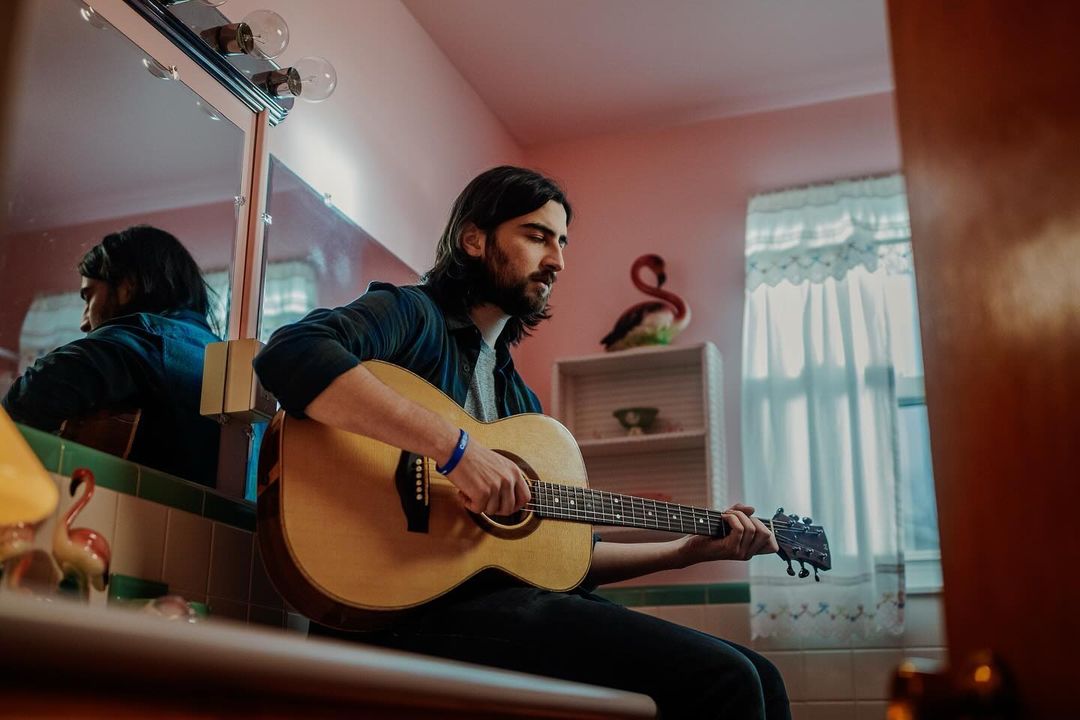 A man playing an acoustic guitar indoors, reflected in a mirror, wearing a dark shirt with a decorative flamingo figure in the background.