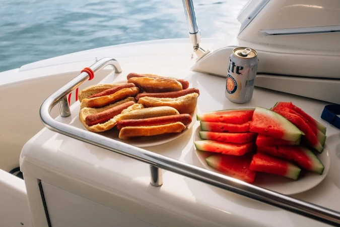 Hot dogs and watermelon slices sit on plates on a boat, accompanied by a can of beer, creating a casual, nautical food spread.