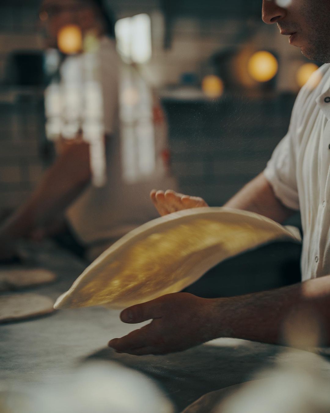 Chef expertly tosses pizza dough at a commercial kitchen counter, preparing for cooking in a restaurant with ambient lighting.