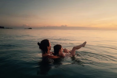 An adult holding a child in calm ocean water during a golden sunset.