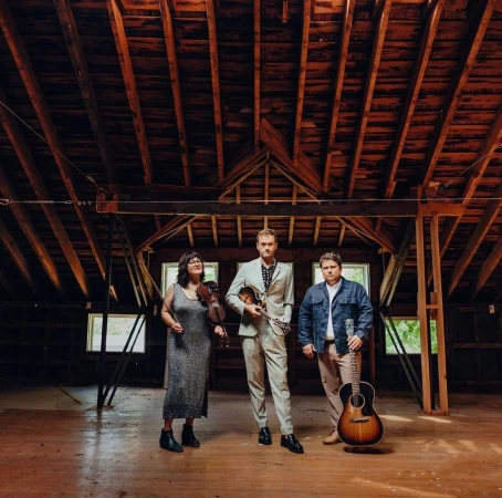 Three musicians posing inside a rustic wooden barn attic with exposed beams, holding a violin, mandolin, and acoustic guitar.