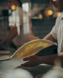 Chef tossing pizza dough in a commercial kitchen with another worker in the blurred background