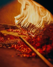 Close-up of wood-fired oven interior with bright flames, burning logs, and glowing embers on a stone hearth.