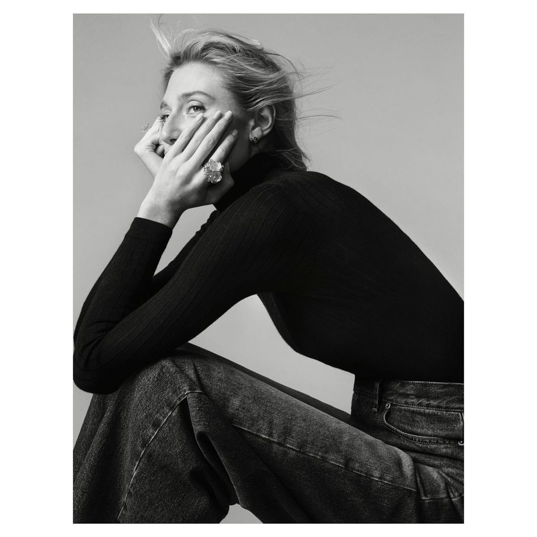 Monochrome portrait of an adult woman with blonde hair wearing a black turtleneck sweater and blue jeans, seated in a photo studio.